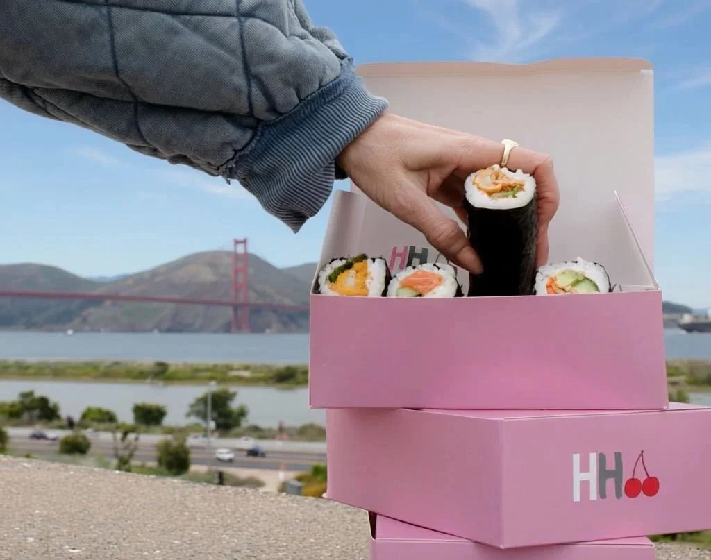 Hand reaching into a box of sushi hand rolls with the Golden Gate Bridge in the background